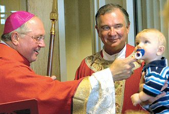 photo of Bishop Morlino, left, greeting seven-month-old Michael Statsick during the blessing of the new buildings at St. Maria Goretti Parish in Madison as Pastor Msgr. Michael Burke looks on